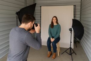 A photographer is taking a portrait of a seated woman in a photo studio set up inside a storage unit.